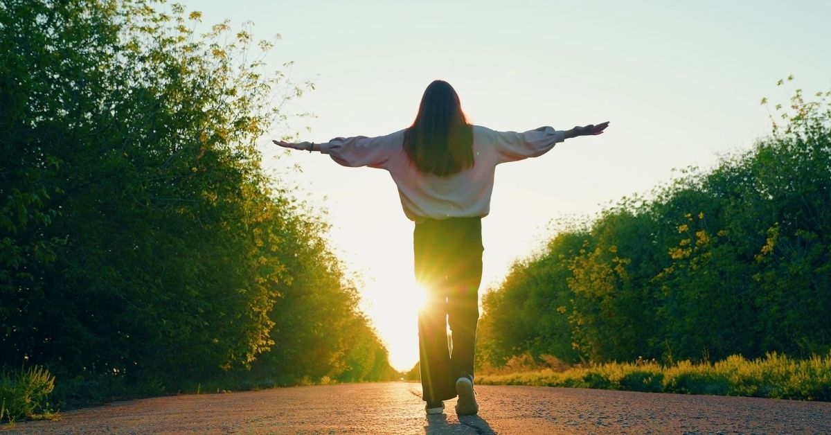 Young woman or teen girl walking on asphalt path toward a sunset or sunrise while raising her hands