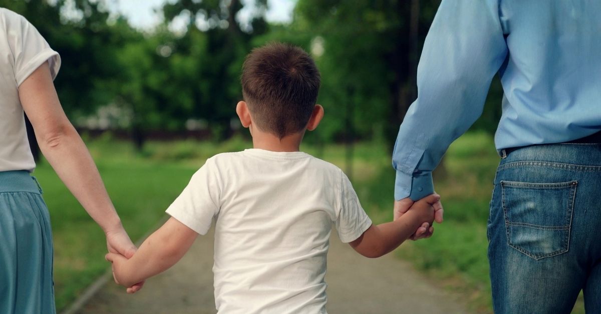 Young boy in white shirt walks and holds hands with mom and dad in park with trees