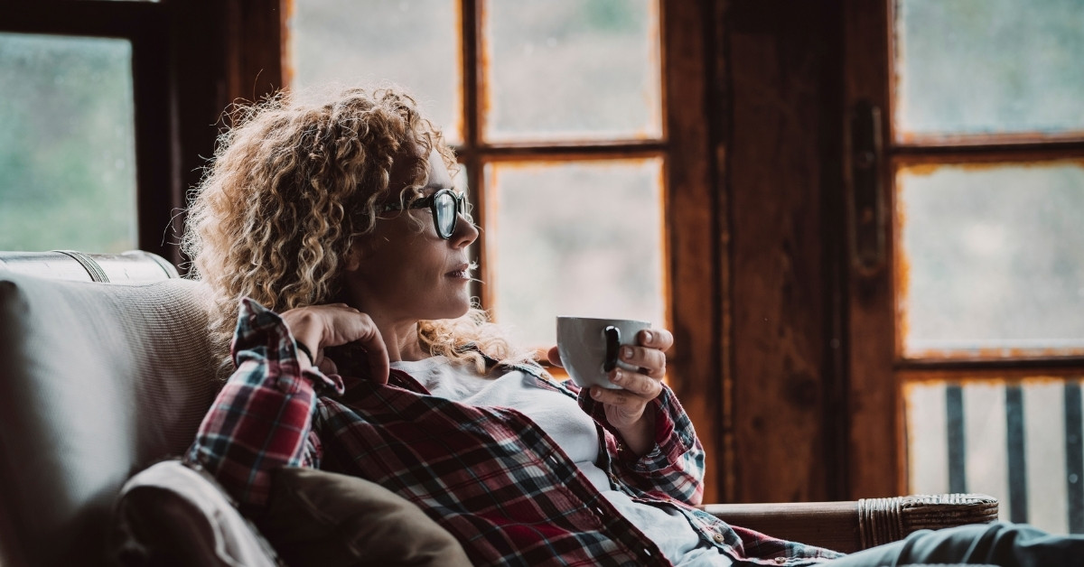 Adult woman with glasses and curly hair, wearing a flannel shirt, sitting and relaxing at home with cup of tea and windows in background, resting in a cozy cabin house for holiday vacation like Thanksgiving or Christmas