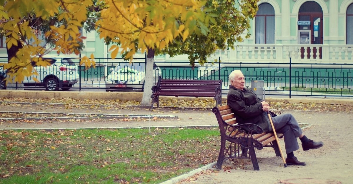 old man wearing a black leather jacket with cane, sitting alone on a park bench where tree leaves have turned yellow in the autumn