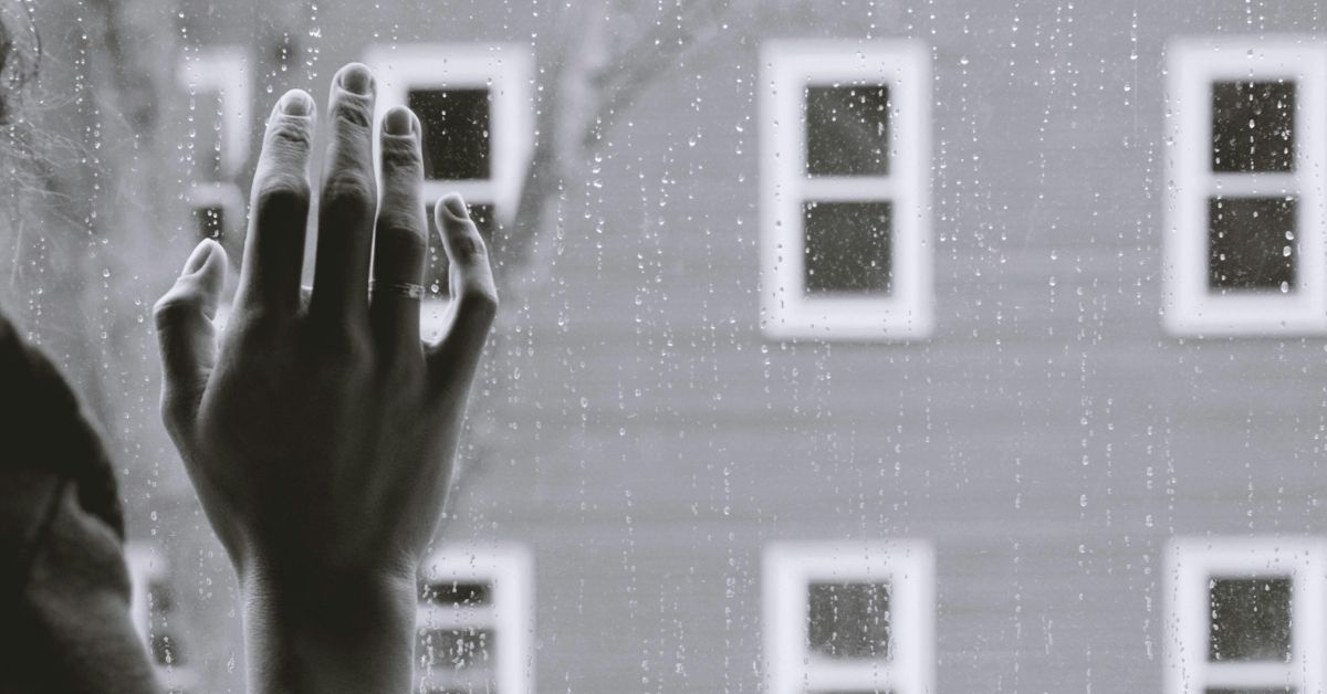 Black and white photo of a woman's hand against window on a rainy day, facing an apartment building outside