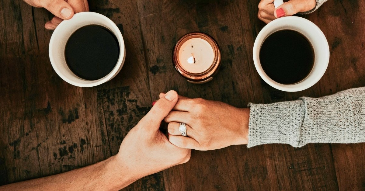 husband and wife holding hands on table next to a lit candle while drinking coffee