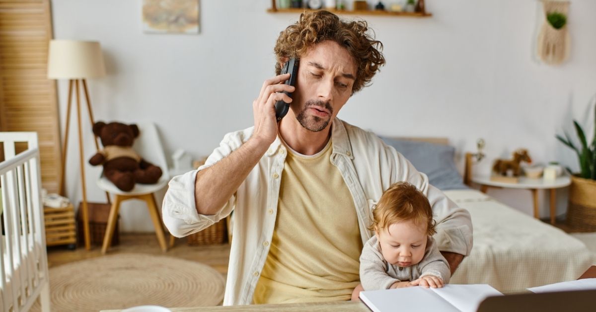 curly-haired, bearded father on the phone, frowning slightly, working at home while caring for his red-headed toddler