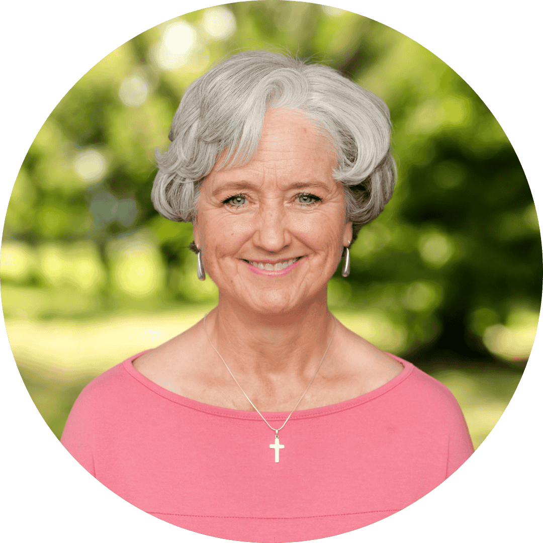 Gray-haired, smiling woman with a short bob haircut, salmon pink shirt, cross necklance, and earrings