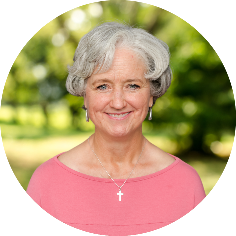 A pleasantly smiling older woman with grey hair in a bright pink shirt in front of a nature background