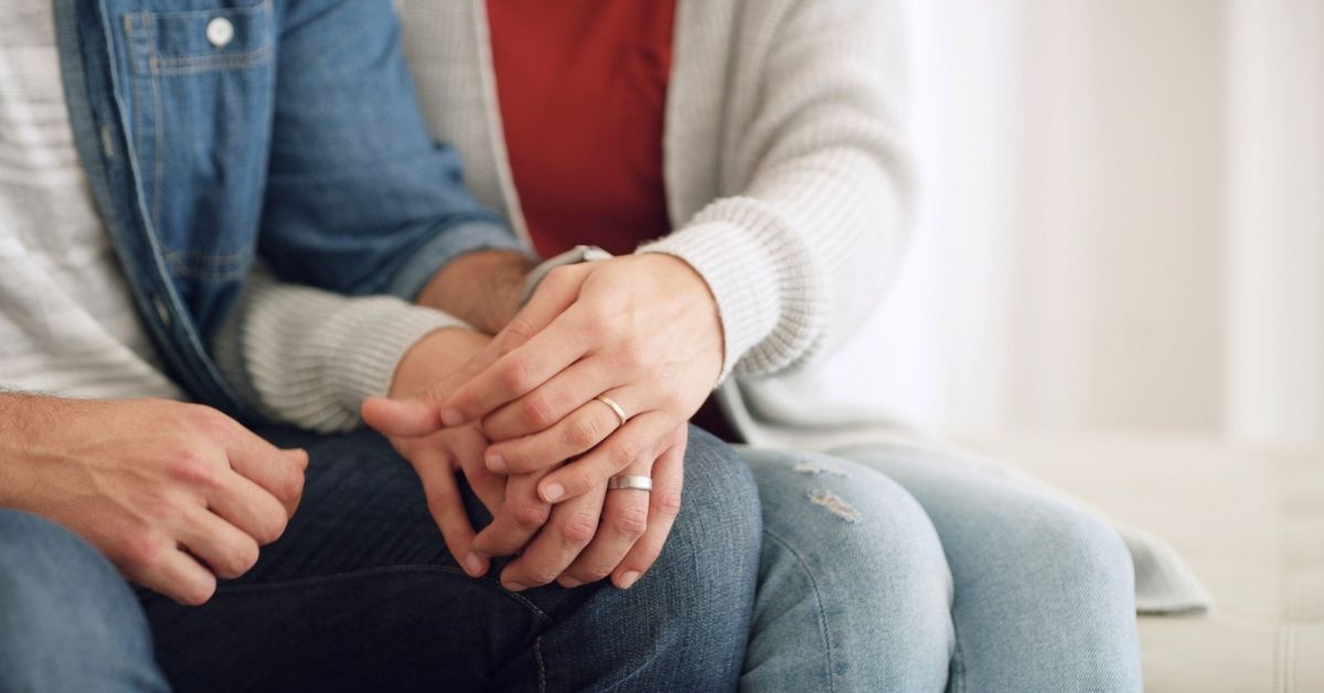 Close-up of a married couple holding hands (with their wedding rings displayed) while sitting on a couch together