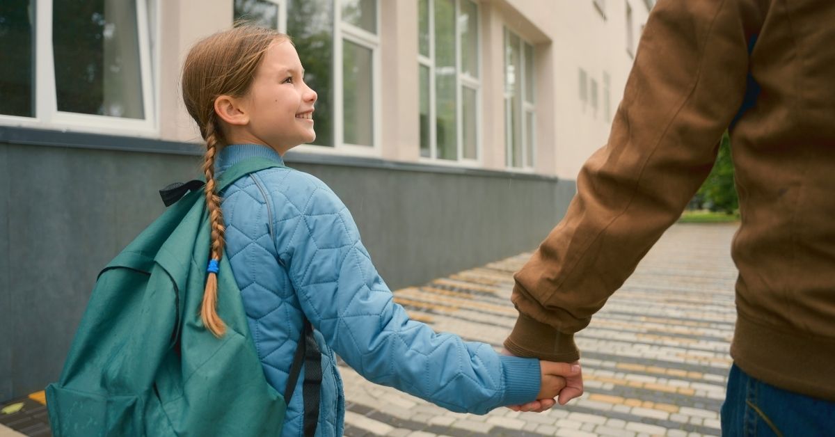 Rear view of young girl in braids wearing a coat and green backpack, walking home from school while holding her father's hand
