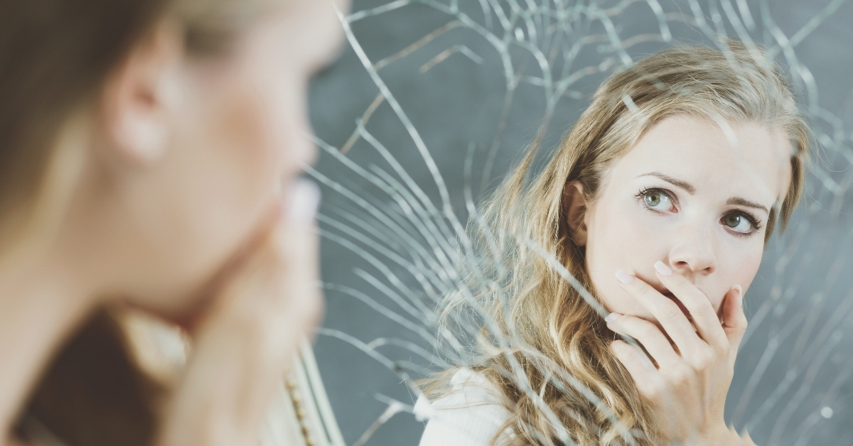 Young blond woman looking in broken cracked mirror while covering her mouth