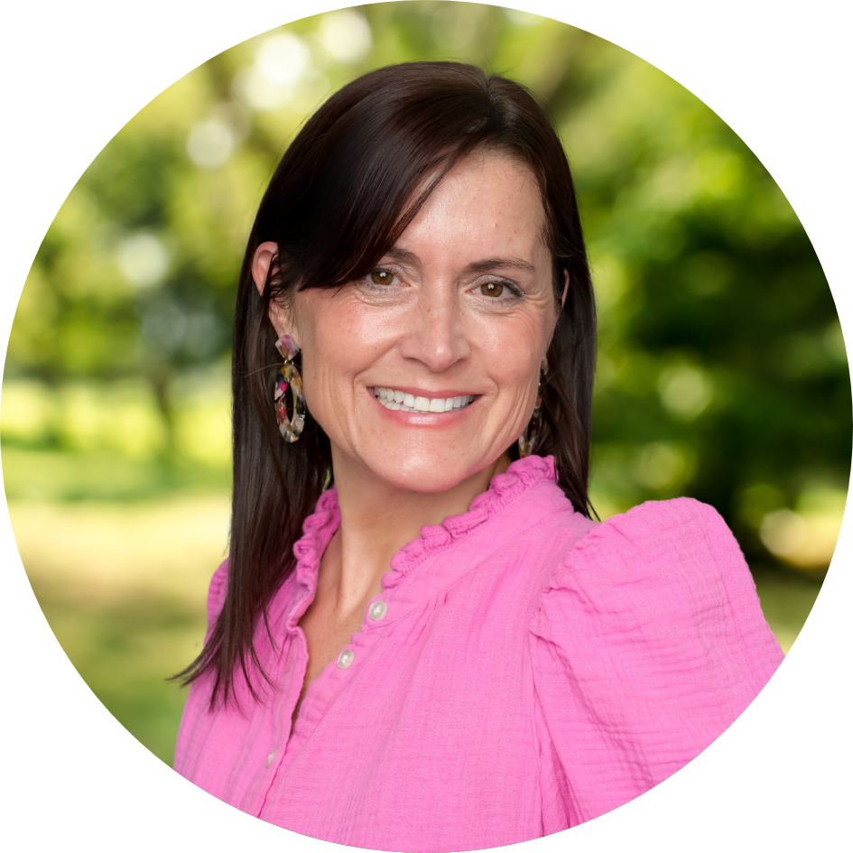 A pleasantly smiling older woman with dark brown hair and a bright pink shirt in front of a nature background