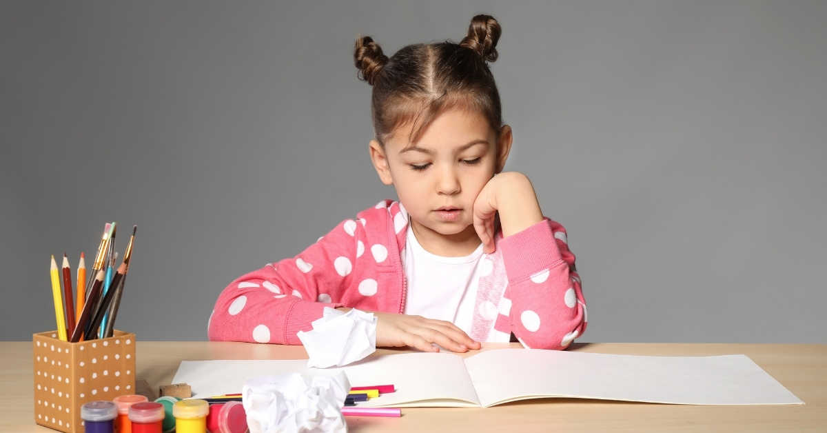 Elementary school girl with brown hair tied up in two buns on the top of her head, wearing a white shirt and pink hoodie with white spots. She is looking at an open, blank book or drawing pad that is partially covered with crumpled balls of paper, colored pencils, and paints.