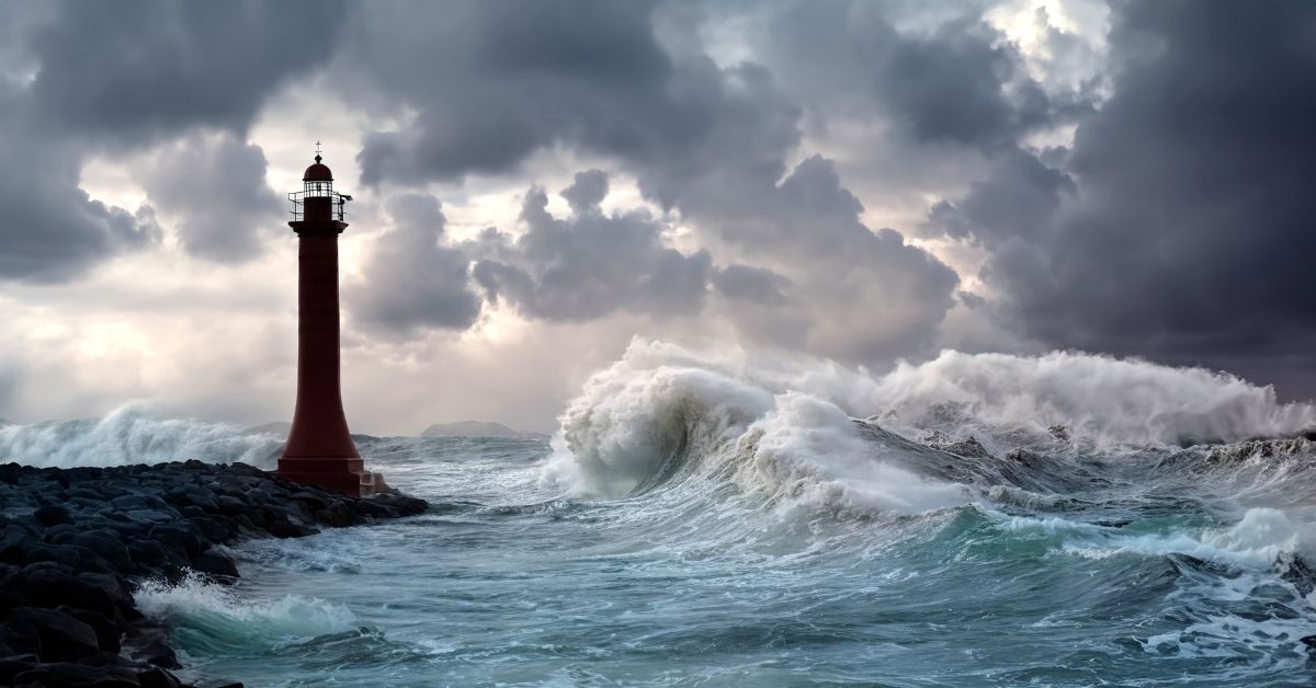 Unlit lighthouse in cloudy stormy weather next to large, crashing rough waves