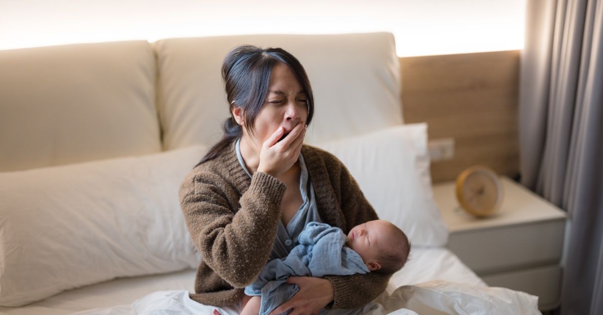 Tired mother with black hair and brown cardigan, yawning while sitting in bed and holding sleeping newborn baby swaddled in blue-gray fabric