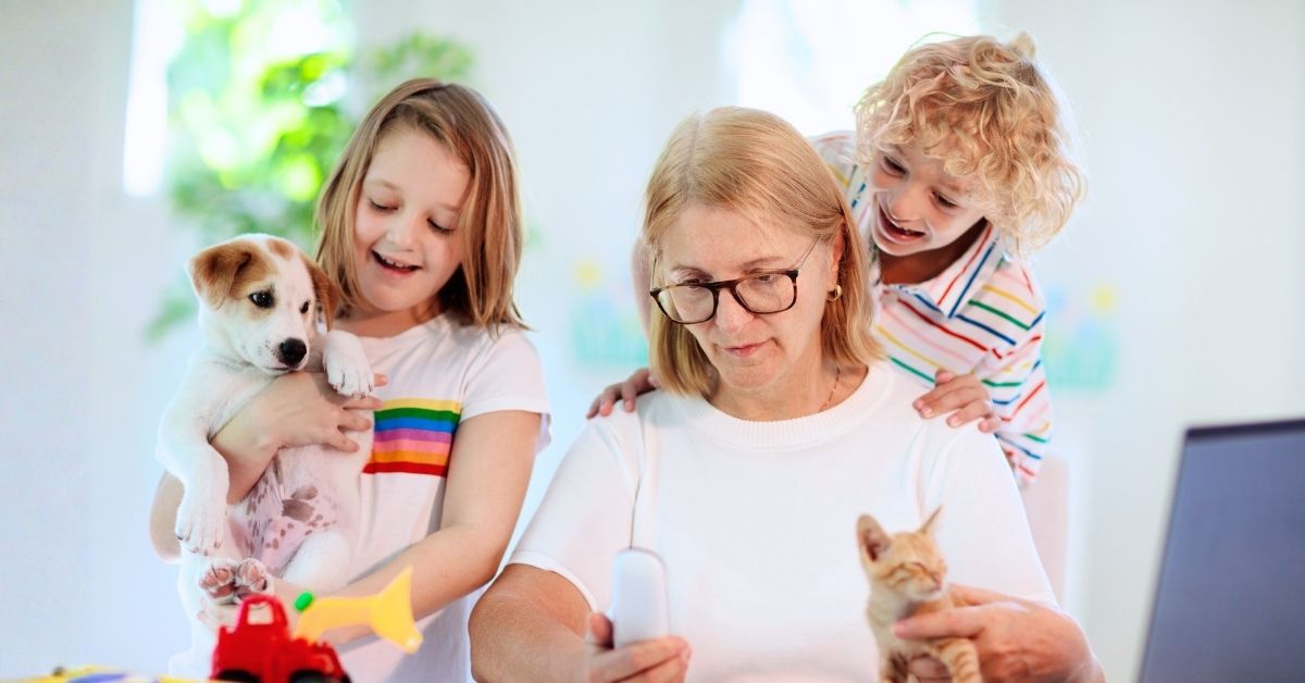 Mom with black glasses is trying to work from home while surrounded by her two younger kids, a dog, and a kitten.