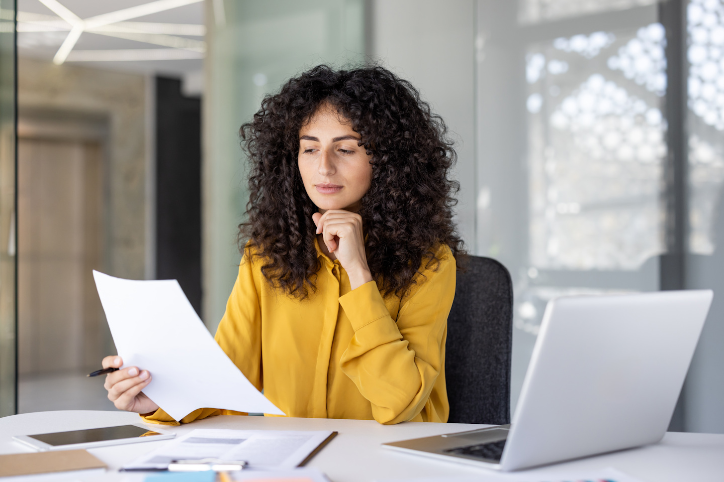 curly-haired brunette woman with hand under her chin, calmly looking at paperwork and laptop at desk