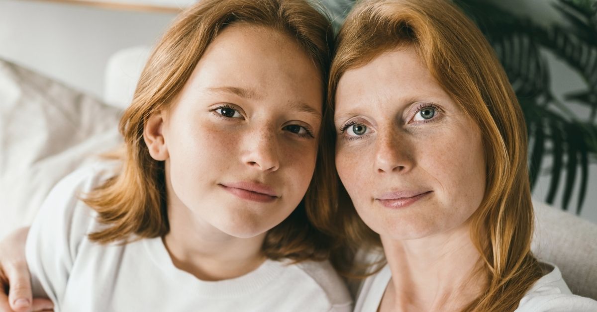 Red-headed mom and teenage daughter, posing cheek to cheek on a sofa