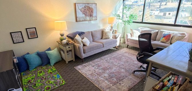 Counseling room with desk, two comfy couches, play therapy toys, and large windows with blinds