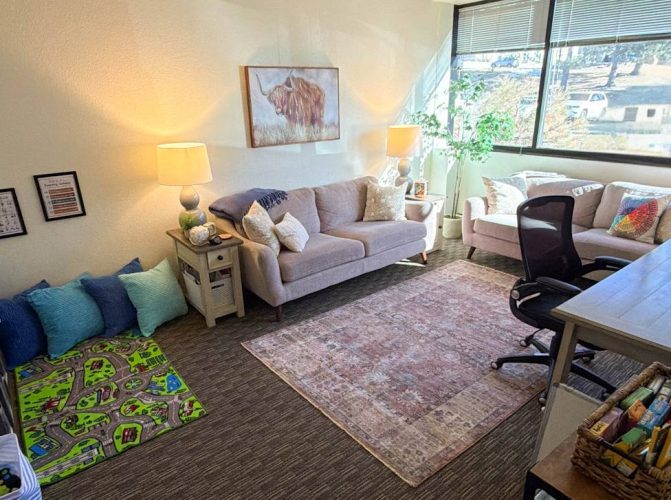 Counseling room with desk, two comfy couches, play therapy toys, and large windows with blinds