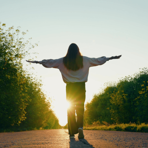 Young woman or teen girl walking on asphalt path toward a sunset or sunrise while raising her hands