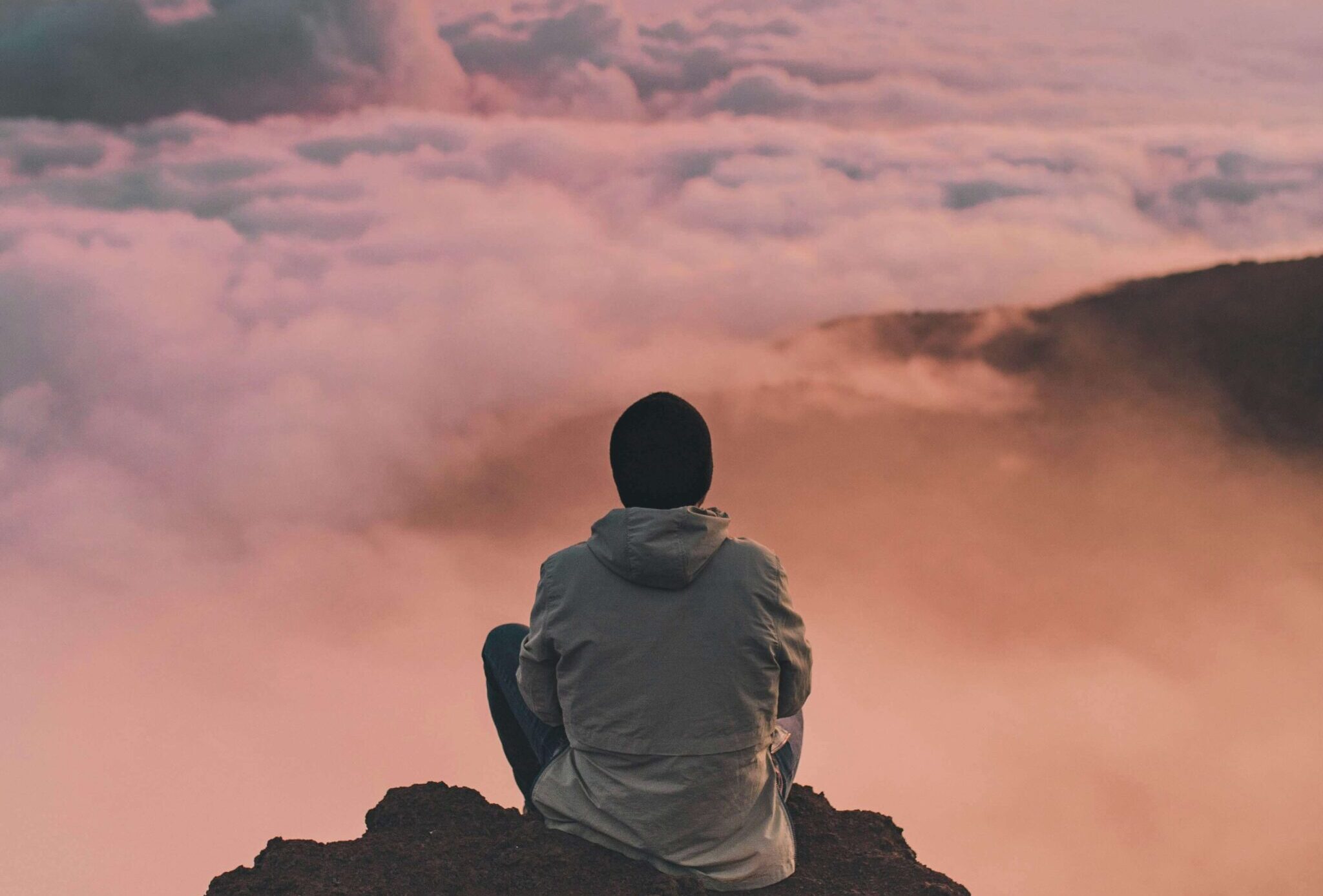A picture of a man sitting on a mountaintop at sunrise, surrounded by clouds.